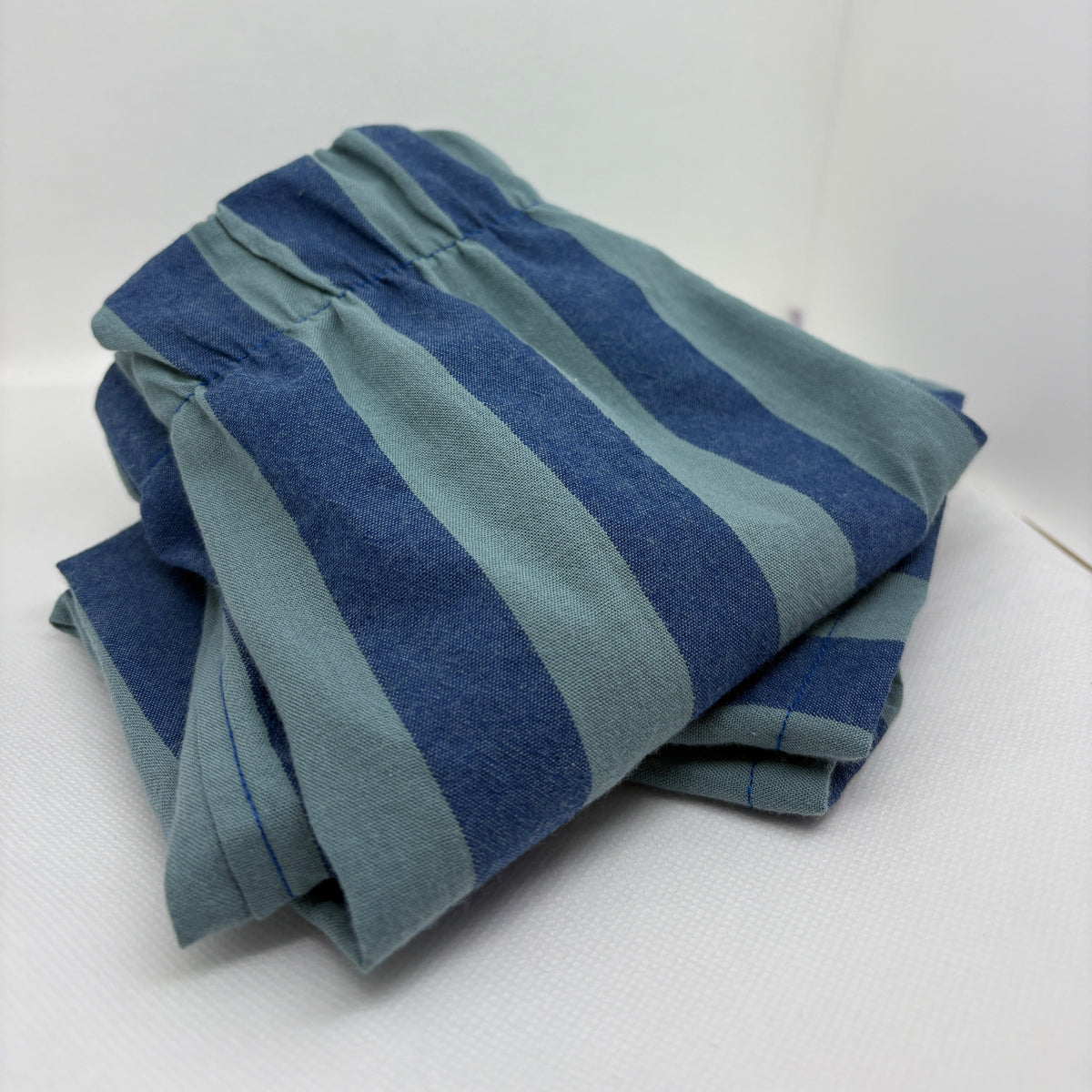 A folded pair of blue and teal striped pants or shorts with an elastic waistband, photographed on a white studio background.